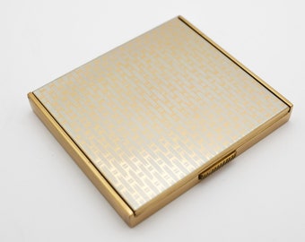 May include: A gold-toned compact mirror with a white surface and a geometric pattern in gold. The mirror has a hinged lid and a small latch.