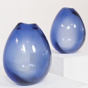 May include: Two blue glass vases with a rounded, teardrop shape. The vases are sitting on a white surface.