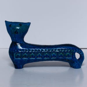 May include: A ceramic cat figurine in shades of blue. The cat has a long body with a decorative pattern of squares and circles. The head is upright with two small holes for eyes. The figurine is approximately 20 cm long.