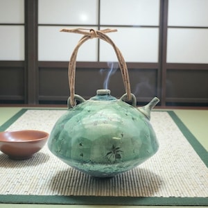 May include: A green ceramic teapot with a textured surface and a woven handle. Steam rises from the spout, indicating hot contents. A small, brown bowl sits nearby on a bamboo mat, suggesting a tea ceremony.