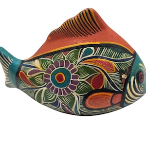 Puede incluir: Figura de pez de cerámica pintada a mano. El pez presenta un cuerpo rojo, aletas turquesas y un diseño floral detallado en verde, morado y amarillo. Es un objeto decorativo.