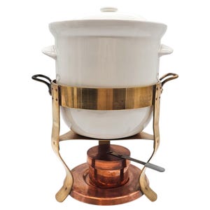May include: A white ceramic fondue pot with a lid, set in a brass and copper stand. The stand has curved legs and a circular base. A small copper burner sits below the pot, with a small metal tool.