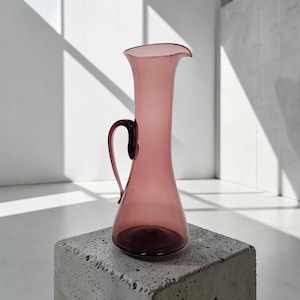 May include: A tall, translucent mauve glass pitcher with a slender neck, a flared rim, and a curved handle attached to the side. The vessel features a smooth, rounded base that tapers upward into a narrow body, showcasing a consistent pinkish-purple hue throughout the glass structure.