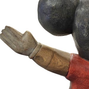 K&ouml;nnte beinhalten: Eine geschnitzte Holzfigur mit erhobener Hand. Der Arm ist braun mit einem roten Hemd und einem schwarzen Kopf. Die Hand ist hellbraun mit sichtbarer Holzmaserung.