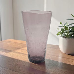 May include: A tall, tapered glass vase in a muted mauve color. The glass features a distinct crackle texture throughout its surface, creating a network of fine lines. The vase has a wide, open rim that narrows toward a solid, circular base.