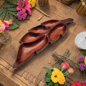 Könnte beinhalten: Eine dunkelbraune, blattförmige Servierplatte aus Holz mit fünf Fächern. Das Tablett ist aus poliertem Holz gefertigt und von bunten Blumen und einer strukturierten Oberfläche umgeben.