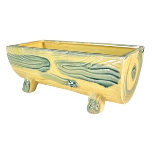 May include: A yellow ceramic planter shaped like a log with green wood grain details. The planter has four short legs and is perfect for displaying plants.