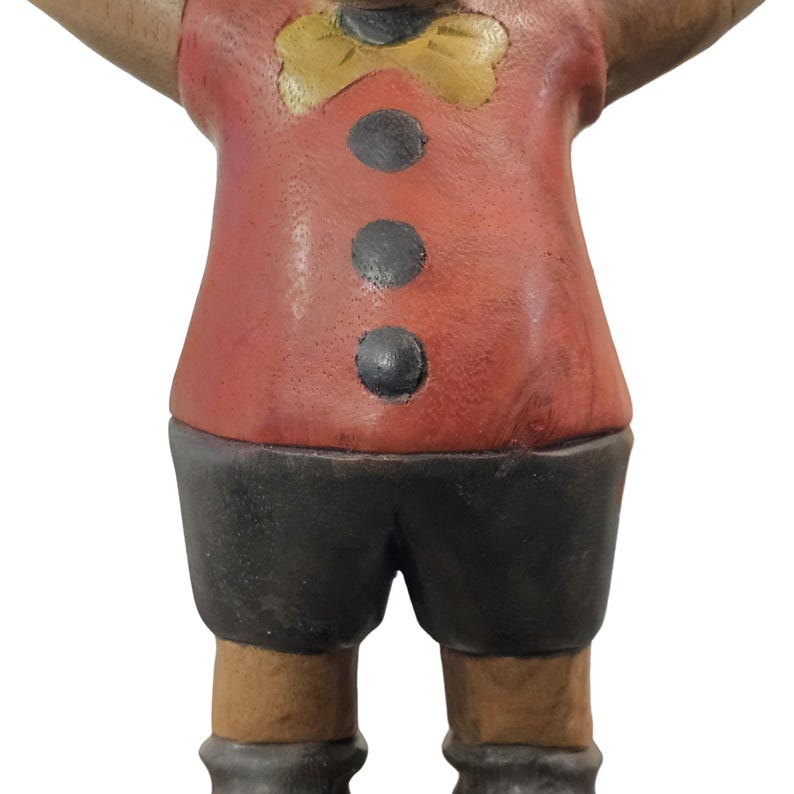 K&ouml;nnte beinhalten: Eine Holzfigur einer Figur, die ein rotes Hemd mit drei schwarzen Kn&ouml;pfen, schwarze Shorts und eine gelbe Fliege tr&auml;gt. Die Figur hat braune Beine und graue Stiefel. Die Figur steht mit ausgestreckten Armen.