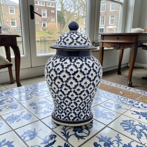 Puede incluir: Un jarrón de cerámica azul y blanco con tapa a juego. El jarrón presenta un patrón repetido de formas florales estilizadas. El jarrón está sobre una superficie de baldosas azules y blancas. Es un elemento decorativo para el hogar.