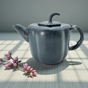 May include: A dark gray ceramic teapot with a curved handle and spout. The lid has a decorative design, and the teapot is next to a sprig of pink flowers. The teapot is on a light-colored surface.