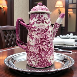 May include: A vintage, tall, ceramic teapot with a lid, handle, and spout. The teapot features a white background with a detailed, repeating pattern in a deep pink color. It sits on a silver-colored, round tray, ready for serving.