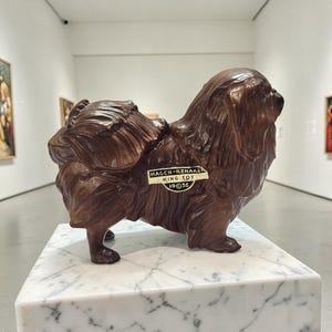 Puede incluir: Una figura de perro de cerámica marrón, etiquetada como "HAGEN-RENAKER MING TOY 19@36", se encuentra sobre un pedestal de mármol blanco. El perro tiene detalles de pelaje y está de perfil. El fondo es una galería.