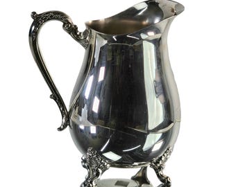 May include: A silver pitcher with a pearlescent finish and three ornate feet. The pitcher has a wide mouth and a curved handle.