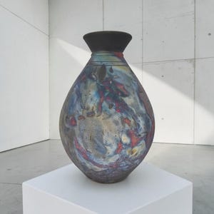 May include: A teardrop-shaped ceramic vase with a wide, flared black rim and a narrow neck. The body features a mottled, multi-colored glaze with swirling patterns of metallic blue, deep red, charcoal gray, and muted gold tones across the surface.