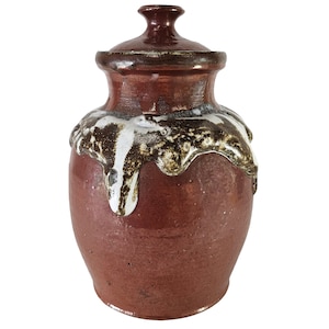 Könnte beinhalten: Eine rote Keramikvase mit Deckel, die mit einer weißen und braunen Glasur verziert ist, die an den Seiten herunterläuft. Die Vase hat einen runden Körper und eine leicht ausgestellte Öffnung.