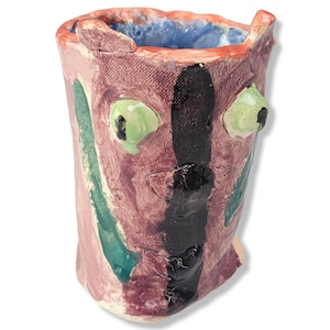 May include: A hand-painted ceramic vase with a whimsical face. The vase is decorated with green, black, and pink paint. It has a textured surface and a wide opening.