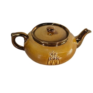 May include: A brown ceramic teapot with a lid and a gold-colored crown and the number 16150 on the side.