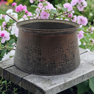 May include: A hammered copper pot with a long handle. The pot has a wide base that tapers to a narrower rim. The copper has a dark, aged patina. The pot is sitting on a weathered wooden surface, with pink and white flowers in the background.