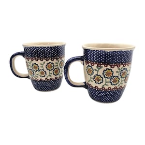May include: Two ceramic mugs with a blue and white floral design. The mugs have a dark blue background with white polka dots and a floral pattern in the center. The handles are a solid blue color.