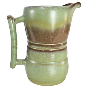 Pode incluir: Uma jarra de cerâmica verde e marrom esmaltada com uma boca larga e uma alça. A jarra tem um aspecto rústico e feito à mão.