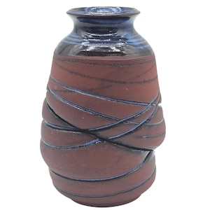 May include: A handcrafted ceramic vase with a dark red body and a dark blue rim. The vase features a unique design of interwoven, raised bands in a lighter red hue, creating a textured surface. The vase is a decorative item.