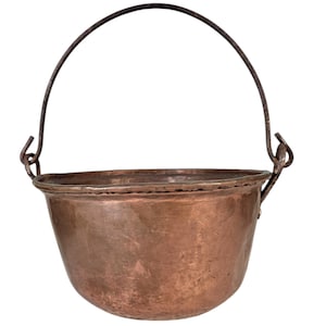 May include: A copper cauldron with a dark metal handle. The cauldron has a rounded base and a slightly flared rim. The handle is attached to the cauldron with metal loops. The copper has a warm, reddish-brown color.