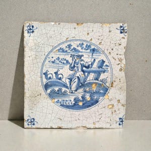Può includere: Antica piastrella di Delft con un'illustrazione blu e bianca di una persona seduta a un tavolo, forse intenta a scrivere. La piastrella presenta un aspetto invecchiato con dettagli dorati e motivi floreali agli angoli.