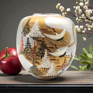 May include: A ceramic vase with a landscape design featuring trees, gold accents, and flying cranes. The vase has a white and gold colour scheme with a round shape. A red fruit is visible on the left side of the image.