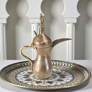 May include: A vintage brass coffee pot with a curved spout and ornate handle sits on a decorative tray. The tray features a gold, white, and black geometric pattern. The background includes white architectural elements.