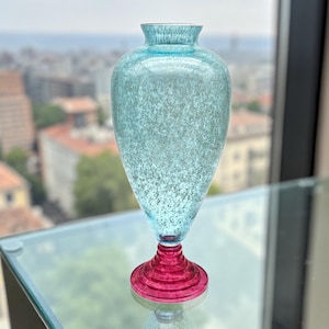 May include: A tall, decorative glass vase with a light blue speckled body and a vibrant red base. The vase has a slender, elongated shape, with a flared top and a rounded base. The base is a deep red color, and the vase is sitting on a glass surface.