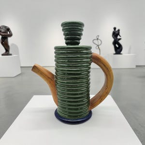 May include: A ceramic teapot with a green, ribbed body, a wooden handle and spout, and a dark blue base. The teapot is displayed in a gallery setting with sculptures in the background.