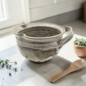 May include: A handmade ceramic mixing bowl with a handle and a spout. The bowl is a neutral color with brown and gray tones. A small wooden spoon and a terracotta pot with a plant are next to the bowl. The bowl is ideal for kitchen use.