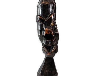 May include: A dark wood sculpture of a stylized human face with a geometric design. The sculpture is carved in a rounded shape and stands on a base.