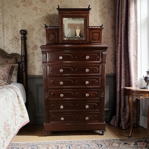 May include: A dark brown antique dresser with a mirror and small cabinets on top. The dresser has six drawers with crystal knobs. The room has floral wallpaper and a bed with a floral quilt.