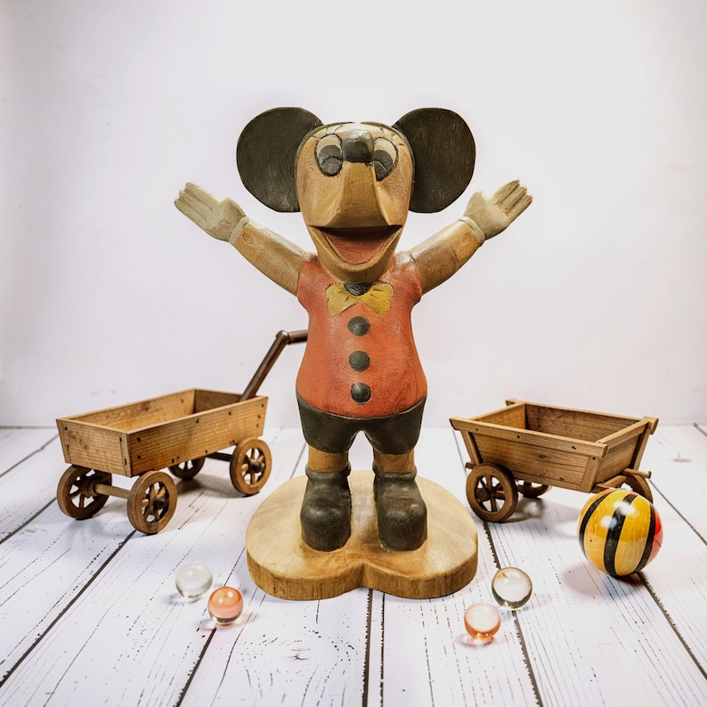 K&ouml;nnte beinhalten: Vintage Holzfigur von Mickey Mouse mit ausgestreckten Armen, rotem Hemd, schwarzen Shorts und Stiefeln. Zwei kleine Holzkarren und Murmeln sind um die Figur platziert. Ein bunter Ball ist ebenfalls vorhanden.
