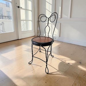 May include: A chair featuring a black wrought iron frame with a decorative scrolled backrest and four twisted metal legs. The circular seat is made of polished brown wood, secured to the metal frame with visible rivets along the outer edge.