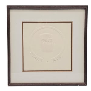 Puede incluir: Obra de arte enmarcada con un marco de madera marrón. La obra presenta un diseño en relieve de color crema dentro de un passe-partout crema. El diseño en relieve incluye un elemento circular con una imagen central y texto en la parte inferior.