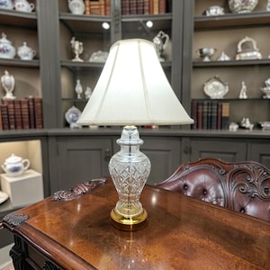 May include: A table lamp featuring a clear, cut-glass base with a diamond-patterned texture and a polished gold-tone metal circular base. The lamp is topped with a tapered, off-white fabric lampshade that emits a soft, warm glow.