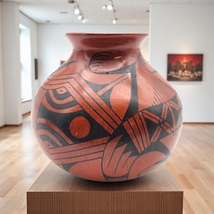 May include: A large, round, terracotta-colored pottery vase with a wide mouth. The vase features a geometric design in black, including lines, circles, and curved shapes. It is displayed on a wooden pedestal in a gallery setting.