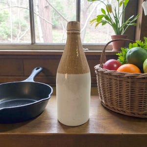 May include: A vintage stoneware bottle with a tan top and white bottom. The bottle is standing on a wooden surface next to a cast iron skillet. A wicker basket filled with fruit sits nearby.