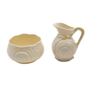 May include: A cream-colored ceramic bowl and pitcher set. The bowl has a scalloped rim and a seashell design. The pitcher has a handle and spout, also with a seashell design. The set is ideal for serving or display.