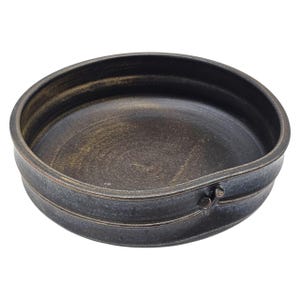 May include: A round, dark brown ceramic bowl with a slightly raised rim and a textured surface. The bowl has a decorative band around the exterior and a small handle-like detail. The interior has a golden hue.