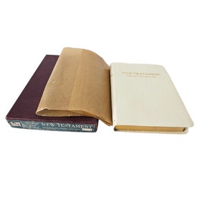 Könnte beinhalten: Ein burgunderfarbenes, ledergebundenes Buch mit dem Titel "World New Testament" und ein weißes, ledergebundenes Buch mit dem Titel "New Testament Red Letter Edition".