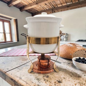 May include: A white ceramic fondue pot with a lid, held within a brass stand featuring two side handles. The stand rests on a circular copper base containing a small burner. The pot is designed for heating and serving food at the table.