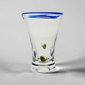 May include: A clear glass tumbler featuring a flared rim with a solid blue glass border. The body of the glass is decorated with small, yellow, fish-shaped glass inclusions embedded within the clear material, creating a subtle aquatic motif.