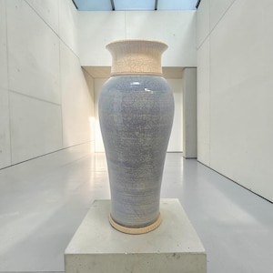 May include: A tall ceramic vase featuring a flared rim and a cylindrical body. The upper neck is a light cream color, while the main body is a soft blue-grey. Both sections display a distinct, fine crackle glaze pattern across the entire surface.