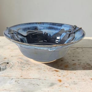 May include: A round ceramic bowl featuring a deep navy blue interior that transitions into a lighter, mottled blue glaze along the wide, flared rim. The exterior surface displays a smooth, solid grey finish with a subtle, glossy sheen throughout.