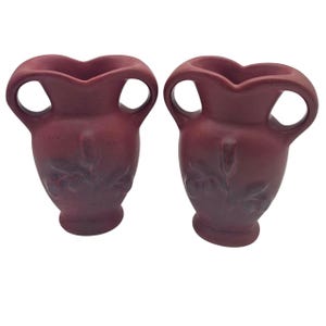 May include: Two maroon-colored vases with a heart-shaped top and two circular handles. The vases have a bulbous base and a floral design on the body. The vases are made of ceramic or pottery.