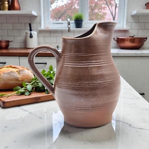May include: A brown ceramic pitcher with a curved handle and a spout. The pitcher has horizontal lines around the body. The pitcher is sitting on a white countertop. A loaf of bread and greenery are in the background.