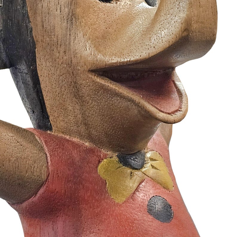 K&ouml;nnte beinhalten: Nahaufnahme einer geschnitzten Holzfigur mit einem l&auml;chelnden Gesichtsausdruck. Die Figur hat braune Haut, schwarzes Haar und tr&auml;gt ein rotes Hemd mit einer gelben Fliege. Ein schwarzer Punkt ist auf dem Hemd zu sehen.